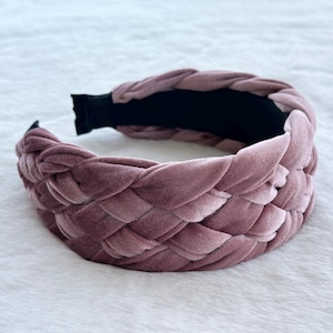 May include: A braided headband in a dusty rose colour. The headband is made of a soft, velvety material and has a black band on the underside. The braided design adds texture to this hair accessory.