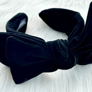 Puede incluir: Una diadema de terciopelo negro con un gran lazo en el centro. La diadema tiene una textura suave y afelpada y un dise&ntilde;o anudado. El lazo es prominente, y el estilo general es elegante y chic.