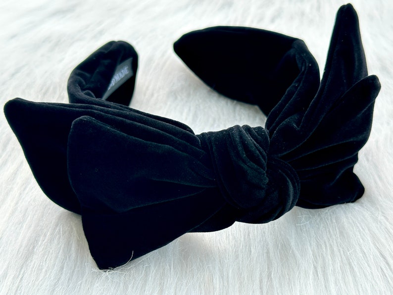 Puede incluir: Una diadema de terciopelo negro con un gran lazo en el centro. La diadema tiene una textura suave y afelpada y un dise&ntilde;o anudado. El lazo es prominente, y el estilo general es elegante y chic.