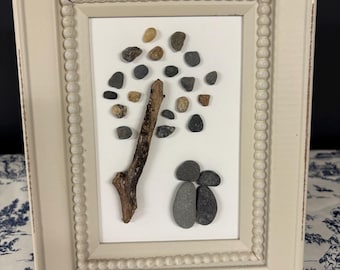 May include: A framed pebble art piece. The artwork features a tree made of a twig and small pebbles in shades of brown, gray, and tan. Two larger gray pebbles represent figures. The frame is off-white with a beaded border.