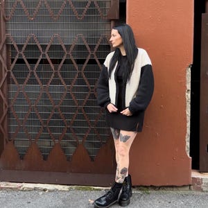 May include: A woman wearing a black and white knitted cardigan, a black mini skirt, and black combat boots. She has tattoos on her legs and is standing in front of a metal gate and a brick wall.