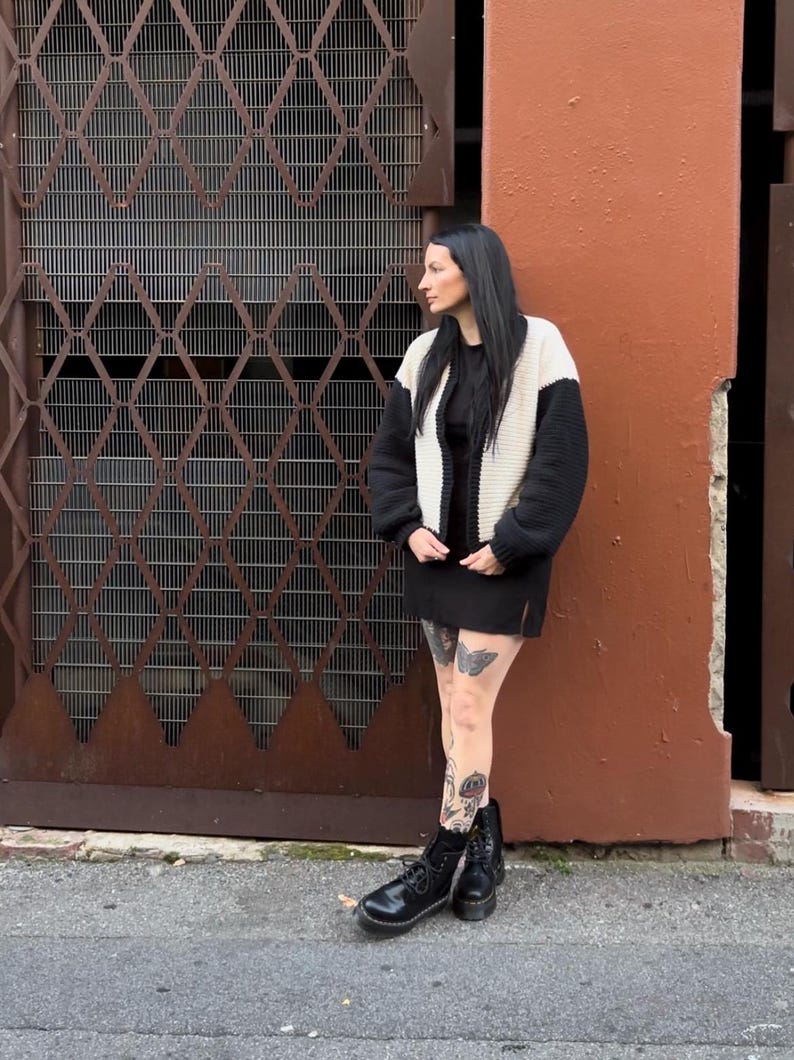 May include: A woman wearing a black and white knitted cardigan, a black mini skirt, and black combat boots. She has tattoos on her legs and is standing in front of a metal gate and a brick wall.