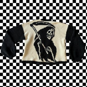 May include: A white knitted sweater with black sleeves and a black and white graphic of a skeleton holding a scythe on the front.