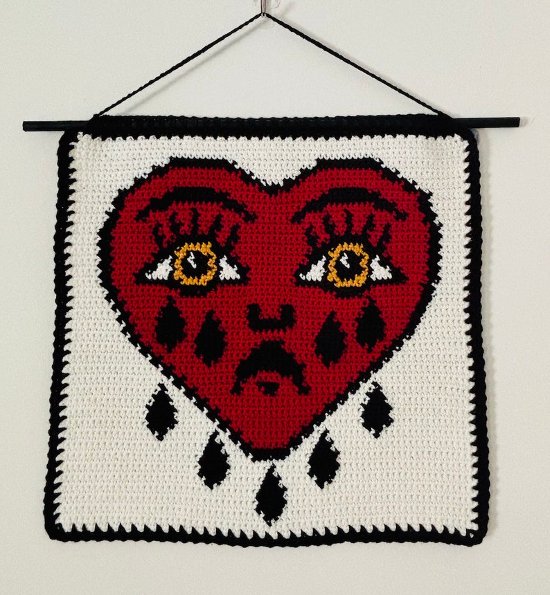 May include: A crocheted wall hanging featuring a red heart with black outlines and yellow eyes. The heart is crying black teardrops. The heart is on a white background with a black border.