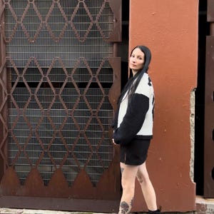 May include: A woman wearing a black and white sweater, a black skirt, and black platform boots. She is standing in front of a brown metal gate and a red wall.