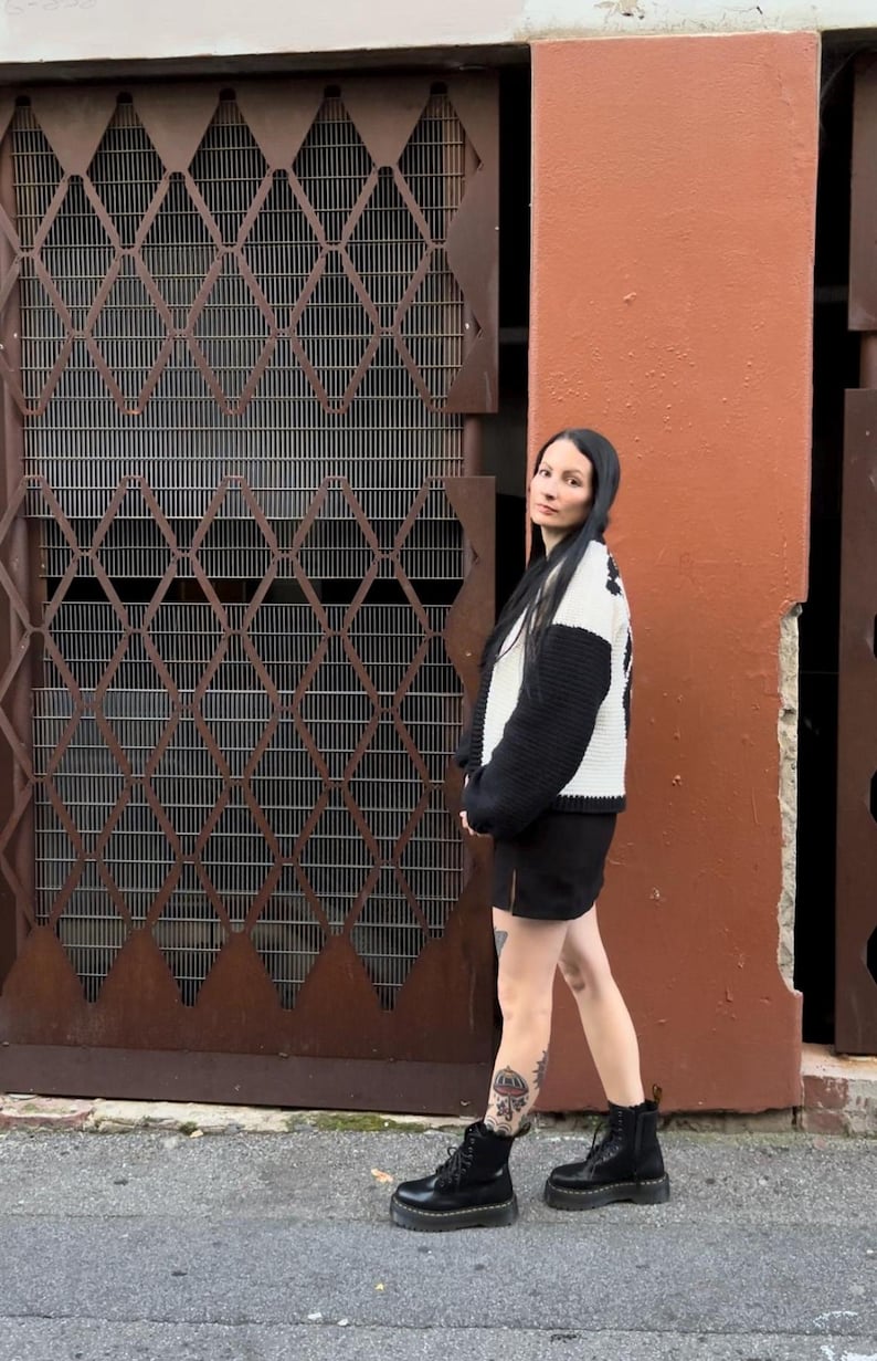 May include: A woman wearing a black and white sweater, a black skirt, and black platform boots. She is standing in front of a brown metal gate and a red wall.