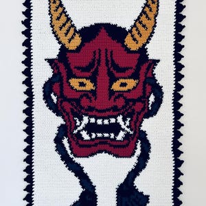 Op de afbeelding: Gehaakte wandhanger met een rood en zwart Oni-masker met gele hoorns. Het masker heeft een woeste uitdrukking met een wijd open mond en scherpe tanden. De achtergrond is wit met een zwarte gehaakte rand.