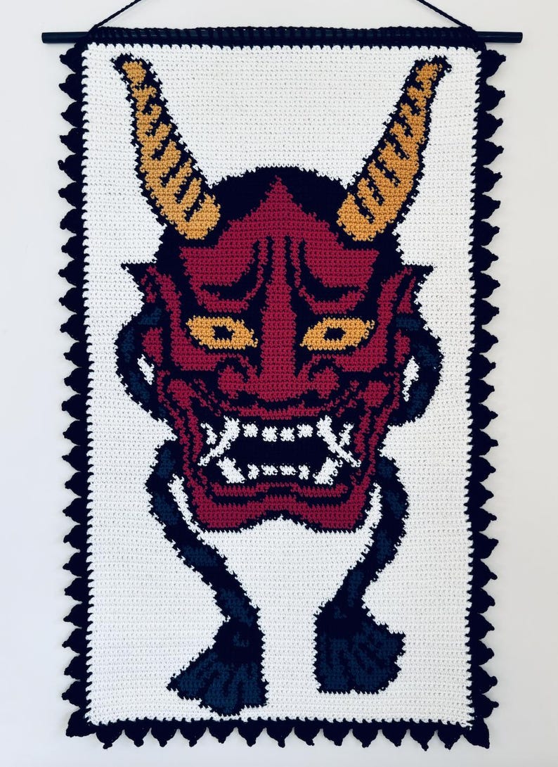Op de afbeelding: Gehaakte wandhanger met een rood en zwart Oni-masker met gele hoorns. Het masker heeft een woeste uitdrukking met een wijd open mond en scherpe tanden. De achtergrond is wit met een zwarte gehaakte rand.