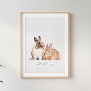 May include: Framed watercolor print featuring two rabbits, one brown and white, the other tan. The print is set against a light background and framed in a light wood frame. The text "Peter & Demeess" is written in cursive below the rabbits.