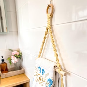 May include: A macrame towel holder with a wooden dowel and ring, hanging on a white tiled wall. A white towel with blue floral embroidery is draped over the dowel. The macrame is a light beige colour.