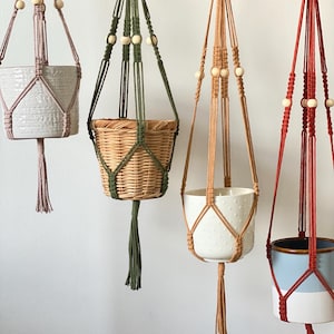 May include: Four macrame plant hangers in different colours. The hangers are made of woven cord and have wooden beads. Each hanger holds a different style of pot, including a woven basket and ceramic pots in white, blue, and pink.
