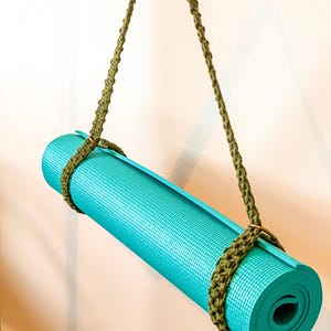 May include: A rolled-up turquoise yoga mat secured with a green macrame strap and hanging from a hook. The macrame strap is looped around the mat and has a braided design. The mat is made of a textured material.