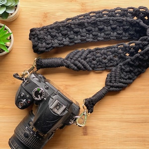 Puede incluir: Correa de cámara de macramé negro con cierres metálicos dorados. La correa está sujeta a una cámara réflex digital negra. La correa está hecha de cuerda de algodón negro tejida. La cámara y la correa están sobre una superficie de madera.