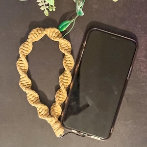 May include: A tan macrame wrist strap next to a black smartphone in a clear case. The wrist strap is made of twisted cord and has a loop design. The phone is on a dark surface with a floral arrangement in the background.