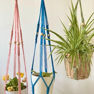 Puede incluir: Tres colgadores de plantas de macramé en rosa, azul y verde. El colgador rosa sostiene una maceta blanca con flores amarillas. El colgador azul tiene una maceta blanca con una planta. El colgador verde tiene una cesta de mimbre con una planta verde.