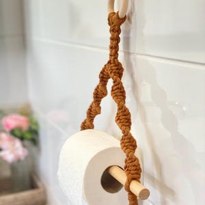 May include: A brown macrame toilet roll holder with a wooden dowel and ring. The holder is suspended from a clear adhesive hook on a white tiled wall. A roll of white toilet paper is in the holder.