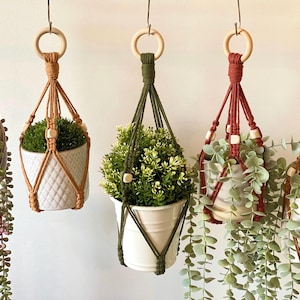 Pode incluir: Cinco suportes de plantas em macramé em várias cores, cada um com um tipo diferente de vegetação. Os suportes são suspensos por anéis de madeira e apresentam contas de madeira. Os vasos são brancos e têm diferentes texturas.