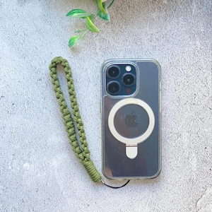 Può includere: Una custodia trasparente per telefono con un anello MagSafe bianco e un cinturino da polso in macramè verde. La custodia del telefono ha una fotocamera a tripla lente. Il cinturino da polso è realizzato in cordoncino verde intrecciato.