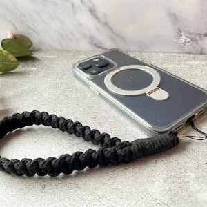 Może przedstawiać: Przezroczyste etui na telefon ze srebrnym pierścieniem i czarnym, plecionym paskiem na nadgarstek. Etui na telefon leży na szarej powierzchni z zielonymi i czerwonymi liśćmi w tle. Pasek na nadgarstek jest wykonany z grubego, tkanego materiału.