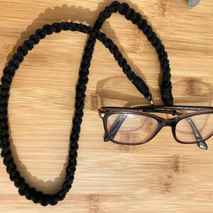 Peut inclure: Porte-lunettes en cordon tressé noir. Le cordon est fait d'un matériau épais et tressé et a un design simple. Le cordon est enroulé autour des branches des lunettes et peut être porté autour du cou.