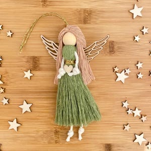 May include: A green yarn angel ornament with wooden wings and a heart. The angel has pink yarn hair and a gold ribbon around its waist. The ornament is hanging from a gold string.