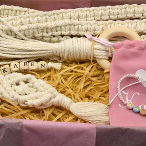 May include: A gift box filled with handmade macrame items, including a keychain, a wall hanging, and a wooden ring. The box also includes a pink pouch with a white heart and a bracelet with colourful beads that spell out the name "Karen".