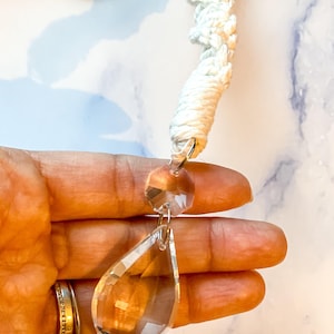 May include: A clear, faceted crystal teardrop pendant hangs from a white macrame cord. A smaller, round crystal bead is above the teardrop. The cord is knotted and looped, creating a decorative element.
