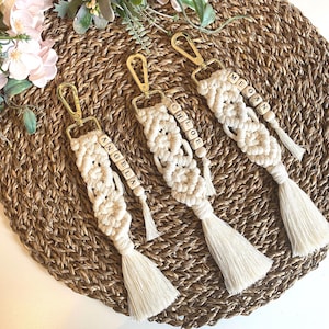 May include: Three white macrame keychains with gold clasps and wooden beads spelling out the names "Angela", "Chloe", and "Megan".