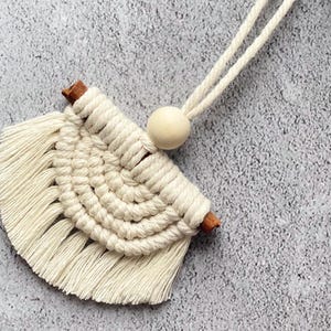 May include: A cream macrame wall hanging in a fan shape. It has a woven design with fringe, a wooden bead, and two small wooden dowels. A cream cord is attached for hanging. The piece is designed for wall decoration.