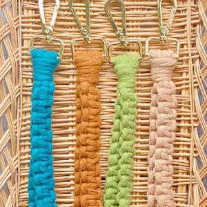 May include: Four macrame keyrings in teal, orange, green, and beige, each with a gold-coloured clasp. The keyrings are displayed in a woven basket, showcasing their handmade quality and vibrant colours.