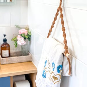 May include: A white embroidered towel with blue floral and gold heart designs hangs from a macrame towel holder. The holder features a wooden dowel and ring, with a brown rope design. The bathroom setting includes a wooden shelf and white tile.