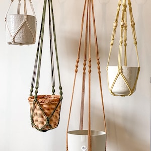 Può includere: Quattro fioriere sospese in vari stili e colori. Una è un cestino intrecciato, un'altra un vaso di ceramica bianca, e altre due sono vasi bianchi con superficie strutturata. I supporti in macramè sono beige, verdi e marroni.