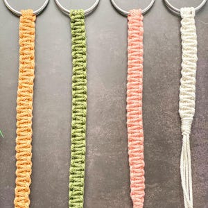 Könnte beinhalten: Vier Makramee-Pflanzenaufhänger in Orange, Grün, Rosa und Weiß. Jeder Aufhänger hat einen silbernen Metallring und ein gewebtes, strukturiertes Design. Die Aufhänger sind zum Aufhängen von Pflanzen gedacht. Die Längen betragen ca. 30 cm.