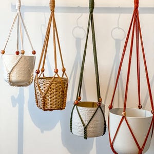 May include: Four hanging planters with different designs. One is white with a dotted pattern, another is woven, and two are white with colored macrame. Each planter is suspended by a macrame hanger with wooden beads, hanging from a metal hook.