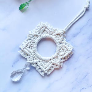 Op de afbeelding: Een handgemaakte, crèmekleurige macramé zonneweerstaan met een stervormig ontwerp. Het cirkelvormige midden is omgeven door een gefranjerde rand. Een helder, druppelvormig kristal hangt aan de onderkant. De zonneweerstaan wordt opgehangen met een geknoopt koord.