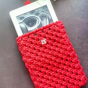 May include: A vibrant red, hand-crocheted e-reader case. The case features a button closure and a textured stitch design. The e-reader screen shows an image. The case is designed to protect the device from damage.