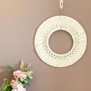 May include: A cream macrame wall decoration, circular in form, hanging from a wall hook. The woven design has a central void and a fringed border. A vase of pink and white flowers is positioned below.
