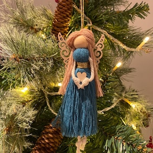Può includere: Un ornamento di angelo in filato blu turchese con una testa di legno e capelli in filato rosa. L'angelo ha ali di legno con un design decorativo e tiene un cuore bianco nelle sue mani.