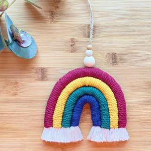 May include: A colourful rainbow wall hanging made of yarn with a burgundy, yellow, green, and blue colour scheme. The rainbow has white tassels at the bottom and a wooden bead at the top.