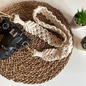 May include: A white crocheted camera strap with gold metal hardware. The strap is lying on a woven brown mat.