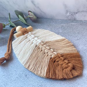 May include: A handmade macrame feather wall hanging in cream, beige and brown. The decorative piece has a wooden bead and dowel, with a knotted cord for hanging. The background includes some greenery.