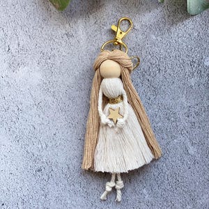Peut inclure: Un porte-clés ange en macramé fait à la main, avec une tête en perle de bois, de longs cheveux beiges et une robe blanche. L'ange tient une petite étoile en bois et a un fermoir doré.