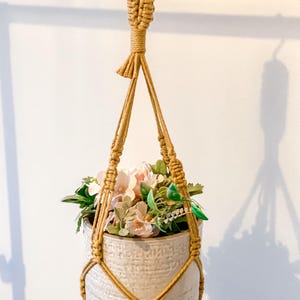 May include: A beige macrame plant hanger with a textured, off-white ceramic pot filled with faux flowers and greenery. The macrame is knotted and woven, creating a decorative and functional piece for displaying plants. The pot is filled with pink and white flowers.