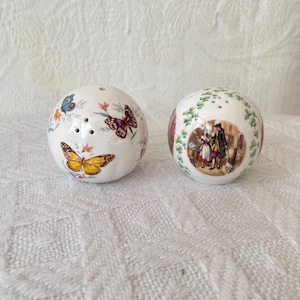 May include: Two spherical white ceramic salt and pepper shakers. One shaker features colourful butterfly and floral illustrations. The other shaker displays a circular scene of two figures in historical attire surrounded by a green vine border.