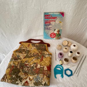 May include: A quilted fabric bag with a brown handle featuring a pastoral print of houses and trees, accompanied by a Mon Tricot Knit & Crochet book, various spools of thread, yarn balls, two metal knitting needles, and blue plastic tools.