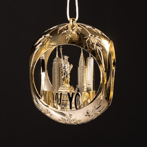 Könnte beinhalten: Ein goldfarbener Weihnachtsschmuck mit einer detaillierten Darstellung der Skyline von New York City, einschließlich der Freiheitsstatue und ikonischer Wolkenkratzer. Das Wort "NEW YORK" ist deutlich sichtbar. Der Schmuck hängt an einem weißen Faden vor schwarzem Hintergrund.