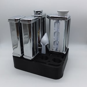 May include: A set of chrome pasta rollers and cutters with a black plastic stand. The stand has slots for each roller and cutter. The rollers and cutters are labelled with the type of pasta they make.