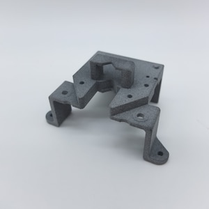 May include: A grey 3D printed plastic part with a rectangular base and a raised section with a hole in the centre.