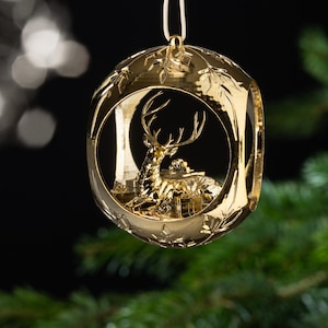 May include: A gold-colored Christmas ornament with a deer and presents inside. The ornament is round with a decorative leaf pattern around the edges. The deer is in a forest setting, and the ornament hangs from a white string.