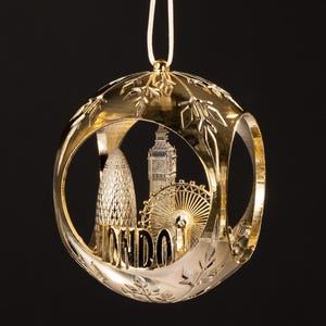 Könnte beinhalten: Ein goldfarbener Weihnachtsschmuck mit London-Motiv. Der Schmuck zeigt Ausschnitte des Gherkin, des Big Ben und des London Eye, mit dem Wort "LONDON" in der Mitte. Er hängt an einer weißen Schnur.